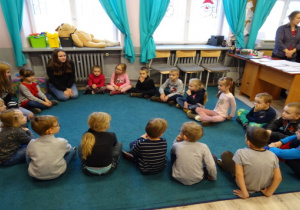 Dzieci siedzą z nauczycielką w klasie, w kole na niebieskiej wykładzinie.